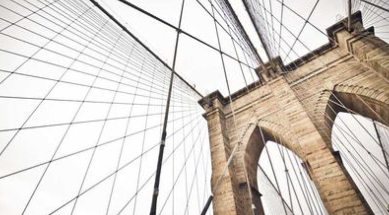 Low-angle view of the Brooklyn Bridge stone tower and suspension cables against a bright sky